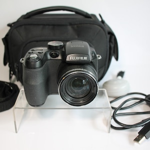 May include: A black Fujifilm digital camera with a prominent lens, featuring a top-mounted flash and control buttons. Included are a black camera bag, a black neck strap, a grey circular lens cap, and a black USB cable for data transfer and charging.