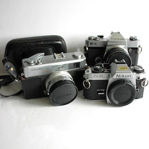 May include: Three vintage film cameras, a silver Minolta, a silver Topcon IC-1, and a silver Nikon FG-20. The cameras are on a white surface.