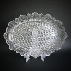 Puede incluir: Bandeja ovalada de vidrio transparente con un centro con diseño de diamantes y un borde con forma de hojas. La bandeja está sostenida por un soporte de plástico transparente, sobre un fondo negro.