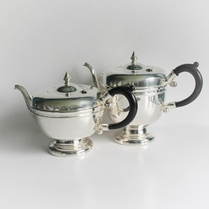 Puede incluir: Dos teteras plateadas con asas negras. Las teteras tienen una superficie brillante y reflectante y están colocadas sobre un fondo blanco. Las asas son curvas y las tapas tienen remates decorativos. Las teteras son de diferentes tamaños.
