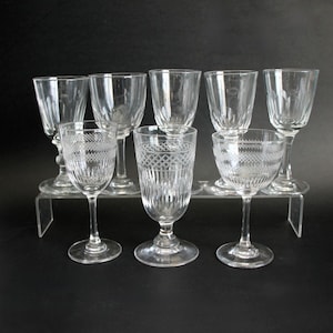 May include: A collection of eight clear glass goblets and glasses. The glasses are arranged on a clear acrylic display stand. Some glasses have etched patterns, while others are plain. The glasses are of varying heights and shapes.