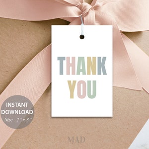 May include: A white gift tag with the words "THANK YOU" in pastel colours. The tag is 5.08 cm x 7.62 cm and is tied with a white string. The tag is on a brown surface with a pink ribbon.