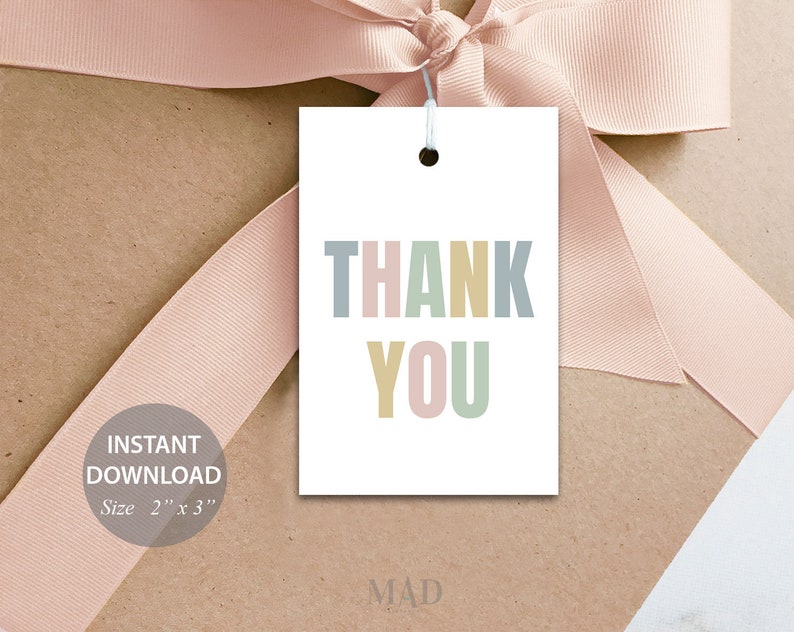 May include: A white gift tag with the words "THANK YOU" in pastel colours. The tag is 5.08 cm x 7.62 cm and is tied with a white string. The tag is on a brown surface with a pink ribbon.