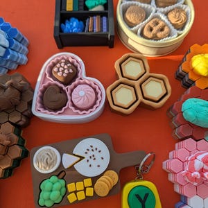 May include: A collection of miniature food and decorative items, including a small bookshelf, a heart-shaped box of chocolates, a round tin of cookies, and a wooden cutting board with cheese and crackers. A yellow keychain with green and red buttons is also present.