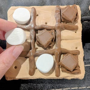 May include: A whimsical tic-tac-toe game set featuring a wooden-textured base with brown game pieces and white marshmallow-shaped game pieces. The game pieces are arranged to resemble a s'more, with chocolate squares and marshmallows.