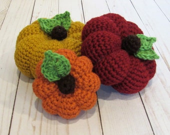 May include: Three crocheted pumpkins in shades of orange, yellow, and red. Each pumpkin has a green leaf and a brown stem. The pumpkins are arranged on a light-colored wooden surface.