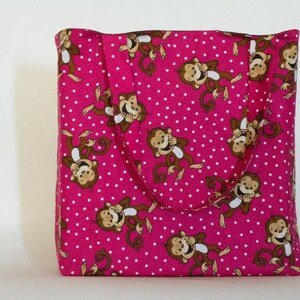 May include: A pink tote bag with a repeating pattern of cartoon monkeys and white polka dots. The monkeys are brown and tan, with their hands covering their eyes. The bag has a handle and is made of fabric.