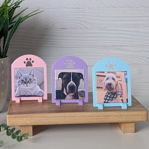以下が含まれることがあります： ピンク、紫、青の3色のペット用フォトフレーム。それぞれ上部に足跡のデザインがあります。フレームには猫と2匹の犬の写真が入っています。フレームは木製のスタンドに飾られています。
