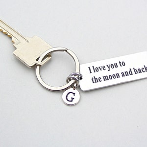 May include: A silver-toned metal keychain featuring a rectangular tag engraved with the text I love you to the moon and back, a circular charm with the letter G, and a standard split ring attached to a silver-colored house key.