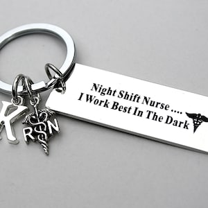May include: A silver-toned metal keychain featuring a rectangular tag engraved with the text "Night Shift Nurse .... I Work Best In The Dark" alongside a caduceus symbol. Two small charms are attached: a letter K and a caduceus with the letters RN.