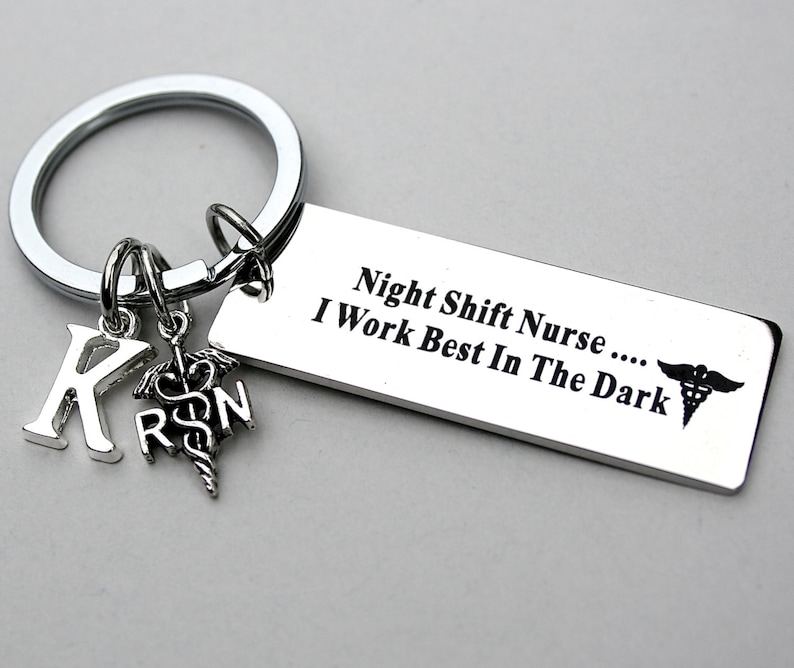 May include: A silver-toned metal keychain featuring a rectangular tag engraved with the text "Night Shift Nurse .... I Work Best In The Dark" alongside a caduceus symbol. Two small charms are attached: a letter K and a caduceus with the letters RN.