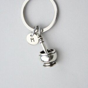 May include: A silver-toned keychain featuring a mortar and pestle charm, with a small round charm displaying the letter "M". The keychain is attached to a silver ring.