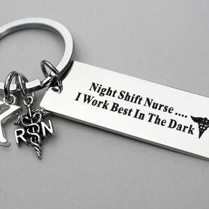 May include: A silver-toned metal keychain featuring a rectangular tag engraved with the text Night Shift Nurse .... I Work Best In The Dark and a caduceus symbol. Attached are two smaller charms: a letter K and a caduceus with the letters RN.