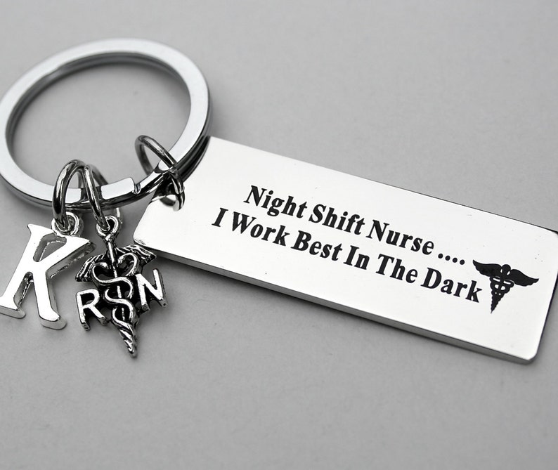 May include: A silver-toned metal keychain featuring a rectangular tag engraved with the text Night Shift Nurse .... I Work Best In The Dark and a caduceus symbol. Attached are two smaller charms: a letter K and a caduceus with the letters RN.