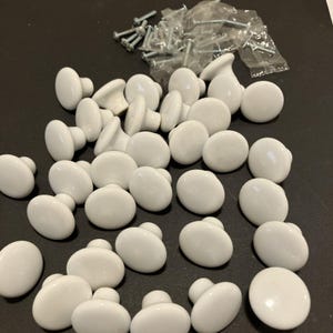 May include: A collection of round, white ceramic cabinet knobs with a smooth, glossy finish. Each knob features a central stem for mounting. Included with the knobs are several small, clear plastic bags containing silver-toned metal mounting screws.