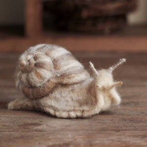 Puede incluir: Una escultura de caracol hecha a mano con fieltro, con una concha marrón y blanca y un cuerpo blanco. El caracol está sentado sobre una superficie de madera.