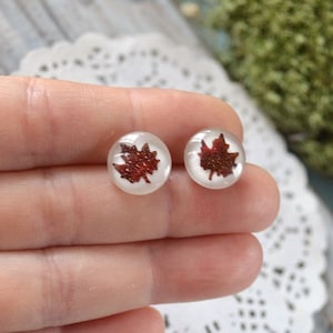 May include: A pair of round stud earrings. Each earring features a red maple leaf design encased in a clear, circular setting. The earrings are held in a hand, resting on a white, lacy surface.