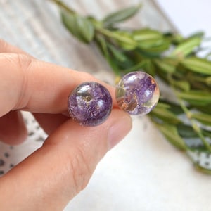 May include: A pair of purple and gold resin earrings with pressed flowers inside. The earrings are round and have a shiny finish.