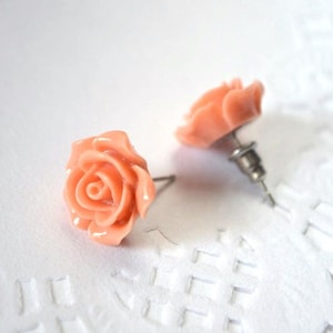 Puede incluir: Un par de pendientes de botón con forma de rosa melocotón. Los pendientes presentan flores de rosa detalladas y están hechos de un material liso. Los postes son plateados. Fondo blanco texturizado.