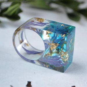May include: A square resin ring with a blue and purple floral design and gold flecks. The ring is clear and has a wide band.