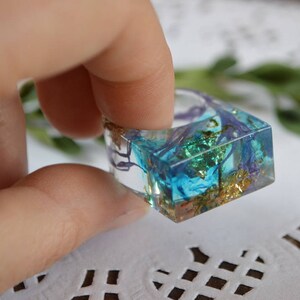 May include: Square resin ring with blue, green, and gold accents. The ring is clear and shows the inclusions inside.
