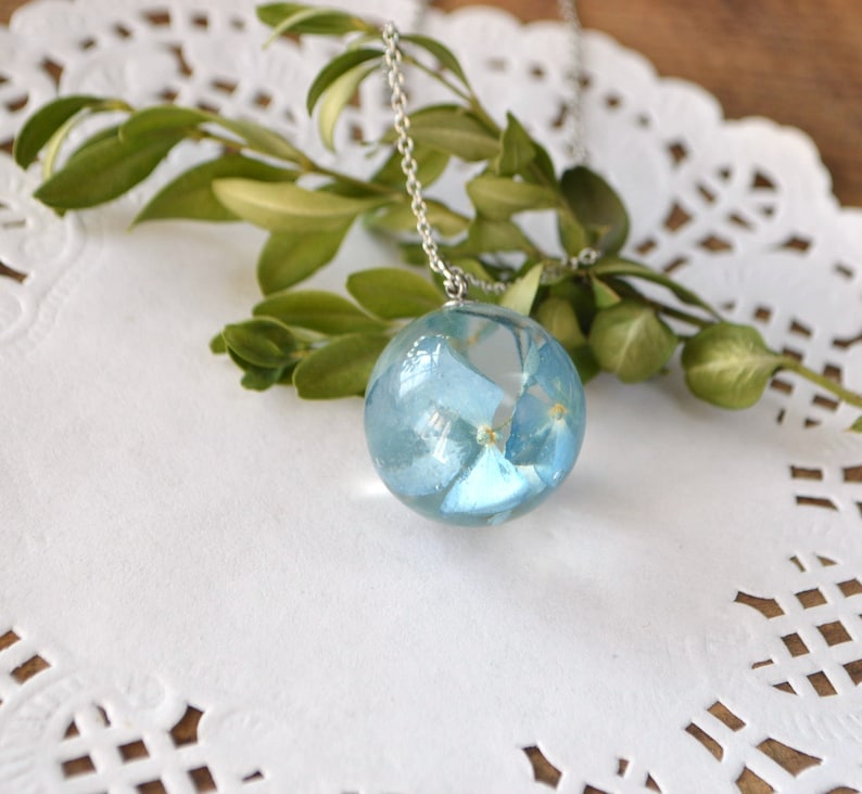 May include: A silver chain necklace with a round pendant featuring blue pressed flowers encased in clear resin.