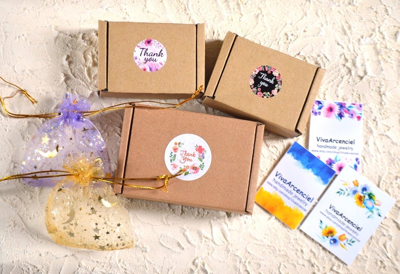 May include: Three brown cardboard boxes with thank you stickers and floral designs. The boxes are tied with gold ribbon. Two clear bags with gold stars and moons are in the foreground. Three business cards with the text "Viva Arcenciel handmade jewelry" are in the background.
