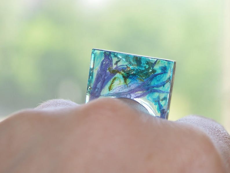 May include: A square ring with a blue and green resin base and embedded dried flowers. The ring is on a person's finger.
