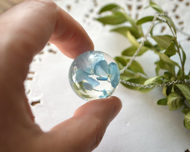 May include: A silver chain necklace with a clear glass pendant containing blue pressed flowers.
