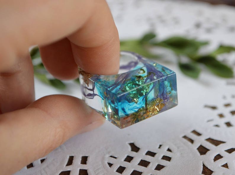 May include: Square resin ring with blue, green, and gold accents. The ring is clear and shows the inclusions inside.