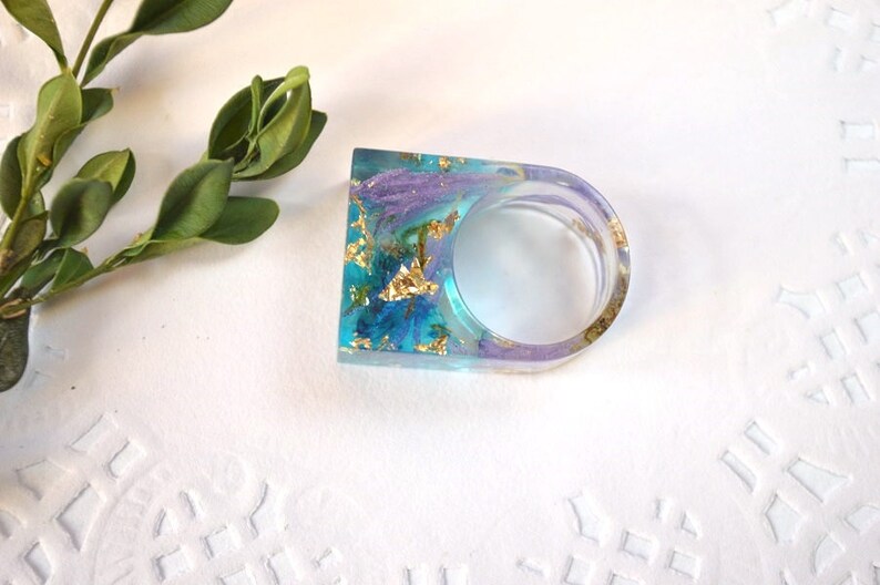 May include: A clear resin ring with a blue and purple gradient, gold flakes, and dried flowers. The ring is on a white background.