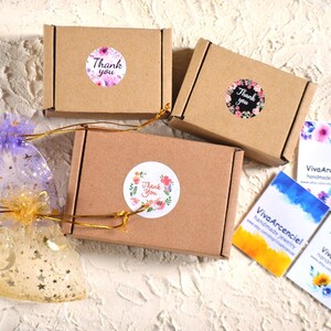 May include: Three brown cardboard boxes with thank you stickers and floral designs. The boxes are tied with gold ribbon. Two clear bags with gold stars and moons are in the foreground. Three business cards with the text "Viva Arcenciel handmade jewelry" are in the background.