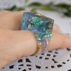 May include: A square-shaped ring with a clear resin base and embedded dried flowers and gold flakes. The ring is a light blue color with hints of green and purple.