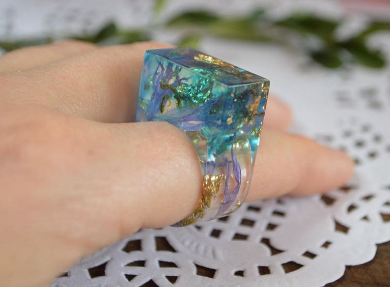 May include: A square-shaped ring with a clear resin base and embedded dried flowers and gold flakes. The ring is a light blue color with hints of green and purple.