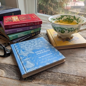 A set of Jane Austen Book Coasters set on a reclaimed wood table. Pride and prejudice in foreground. Mansfield park, Emma, Sense and Sensibility, and Northanger Abbey in the background. And a yellow cover of Persuasion under a fancy Teacup.