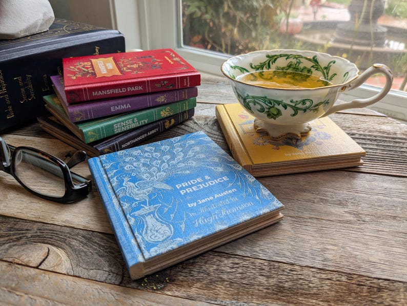 A set of Jane Austen Book Coasters set on a reclaimed wood table. Pride and prejudice in foreground. Mansfield park, Emma, Sense and Sensibility, and Northanger Abbey in the background. And a yellow cover of Persuasion under a fancy Teacup.