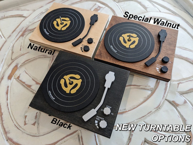 May include: Four miniature wooden record players in different wood finishes: natural, black, special walnut, and a light wood. Each player has a black record with a gold 45 rpm adapter and a white tone arm.
