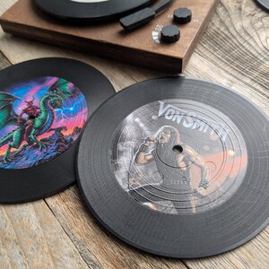 May include: Two black vinyl record coasters. One features a green dragon with red wings and lightning bolts. The other features a black and white image of a person singing with the text "Von Smith" on the record.