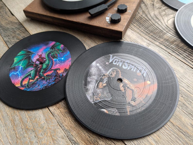 May include: Two black vinyl record coasters. One features a green dragon with red wings and lightning bolts. The other features a black and white image of a person singing with the text "Von Smith" on the record.