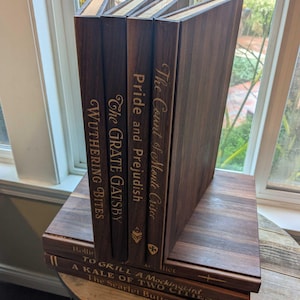 8 Book shaped cutting boards sit in a stack with punny titles on the spine such as Wuthering Bites, the Count of Monte Crisco, and to grill a mockingbird.