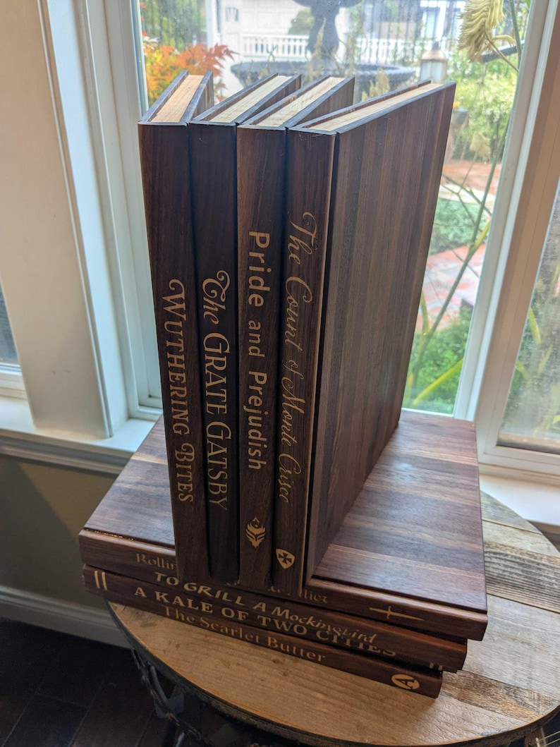 8 Book shaped cutting boards sit in a stack with punny titles on the spine such as Wuthering Bites, the Count of Monte Crisco, and to grill a mockingbird.