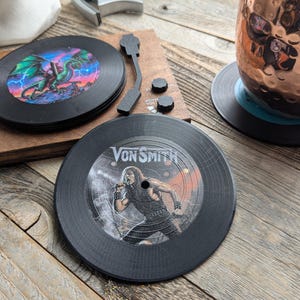 May include: Two black vinyl record coasters. One features a green dragon with lightning bolts and the other features a black and white image of a person singing with the text "Von Smith".