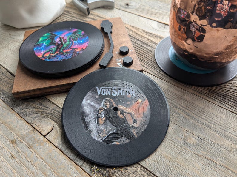 May include: Two black vinyl record coasters. One features a green dragon with lightning bolts and the other features a black and white image of a person singing with the text "Von Smith".