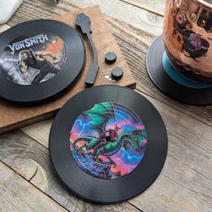 May include: Two black vinyl record coasters. One coaster features a band called "Von Smith" with a person in a black and white outfit. The other coaster features a green dragon with a red figure riding on its back.