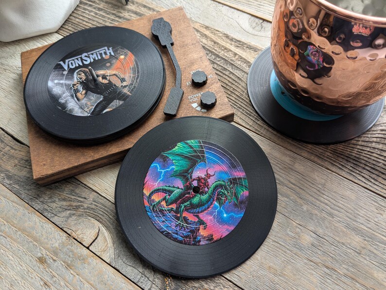 May include: Two black vinyl record coasters. One coaster features a band called "Von Smith" with a person in a black and white outfit. The other coaster features a green dragon with a red figure riding on its back.