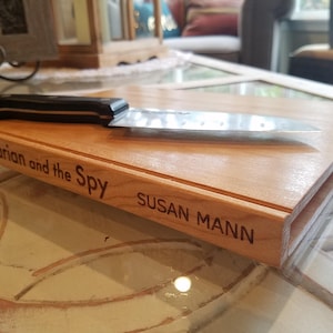 May include: A wooden cutting board with the text "The Librarian and the Spy" and "SUSAN MANN" engraved on the side. A large knife with a black handle rests on the board. The board is light brown.