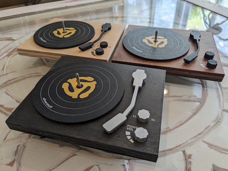 May include: Three wooden record player models with black turntables and gold vinyl record labels. The turntables have a silver tone arm and knobs for power, volume, and speed. The models are on a white surface.