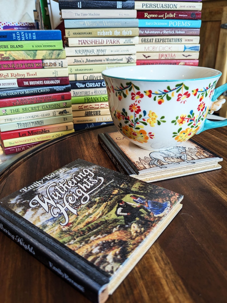 A Wuthering Heights Book wooden book coaster sits in the foreground. Canterbury Tales book coaster sits under a flowery teacup, and a stack of book coasters with the spines showing occupies the background.