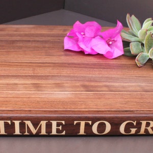 Pu&ograve; includere: Un tagliere in legno marrone con la scritta "A Time To Grill" incisa sul davanti. Il tagliere &egrave; decorato con fiori rosa e piante grasse verdi.