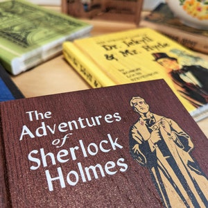 A close up detail of the Adventure of Sherlock Holmes Wooden Book Coaster is in the foreground. Behind you can see the yellow cover of Dr Jeckyll and Mr Hyde, and The green cover of Frankenstein.